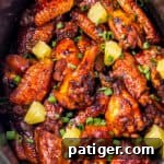 A crockpot packed with pineapple brown sugar soy sauce Hawaiian wings. The chicken wings are topped with chunks of pineapple and chopped scallions.