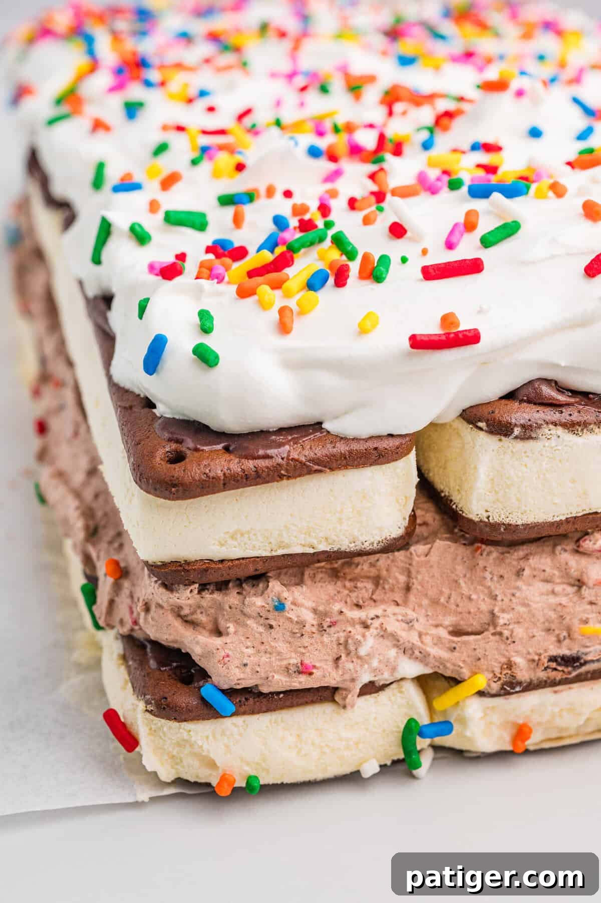 lose-up corner view of a layered ice cream sandwich cake. The cake consists of alternating layers of vanilla ice cream sandwiches and chocolate whipped filling with colorful sprinkles, topped with a thick layer of whipped topping and more rainbow sprinkles.