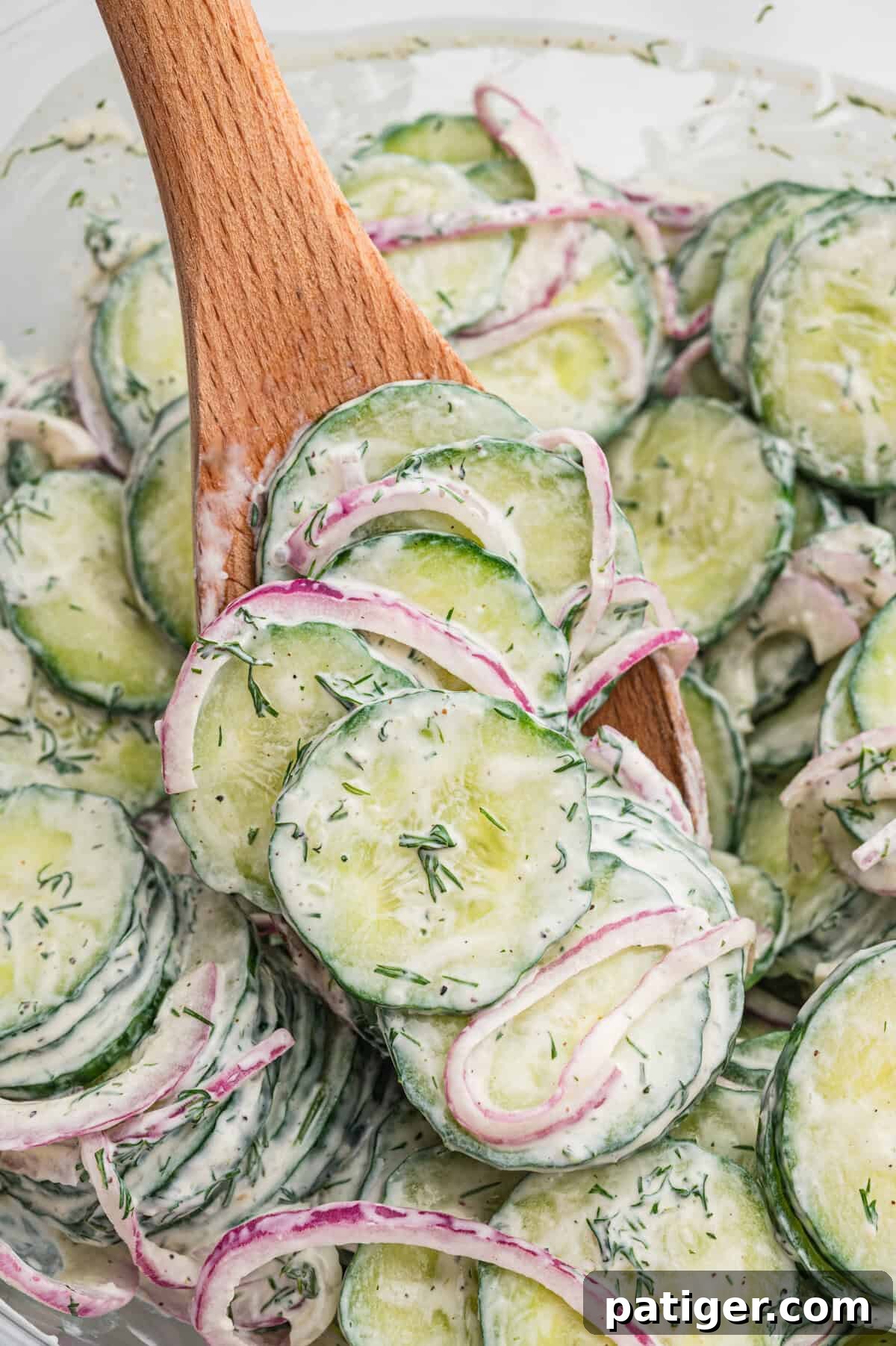 Wooden spoon lifting creamy cucumber salad from a bowl, showing thin cucumber slices, red onion, and specks of dill in a creamy dressing.