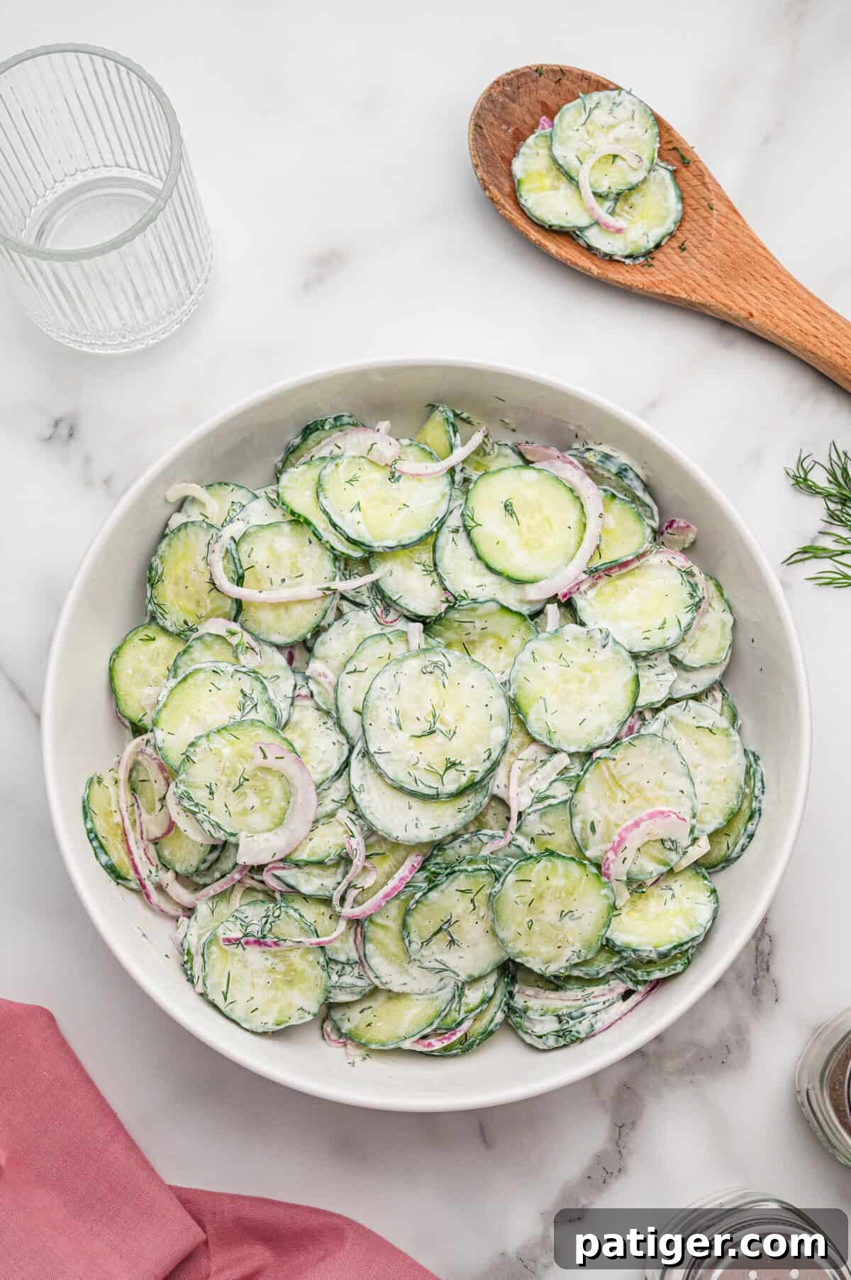 Bowl of creamy cucumber dill salad on a marble surface, with a wooden spoon holding more salad resting beside it.