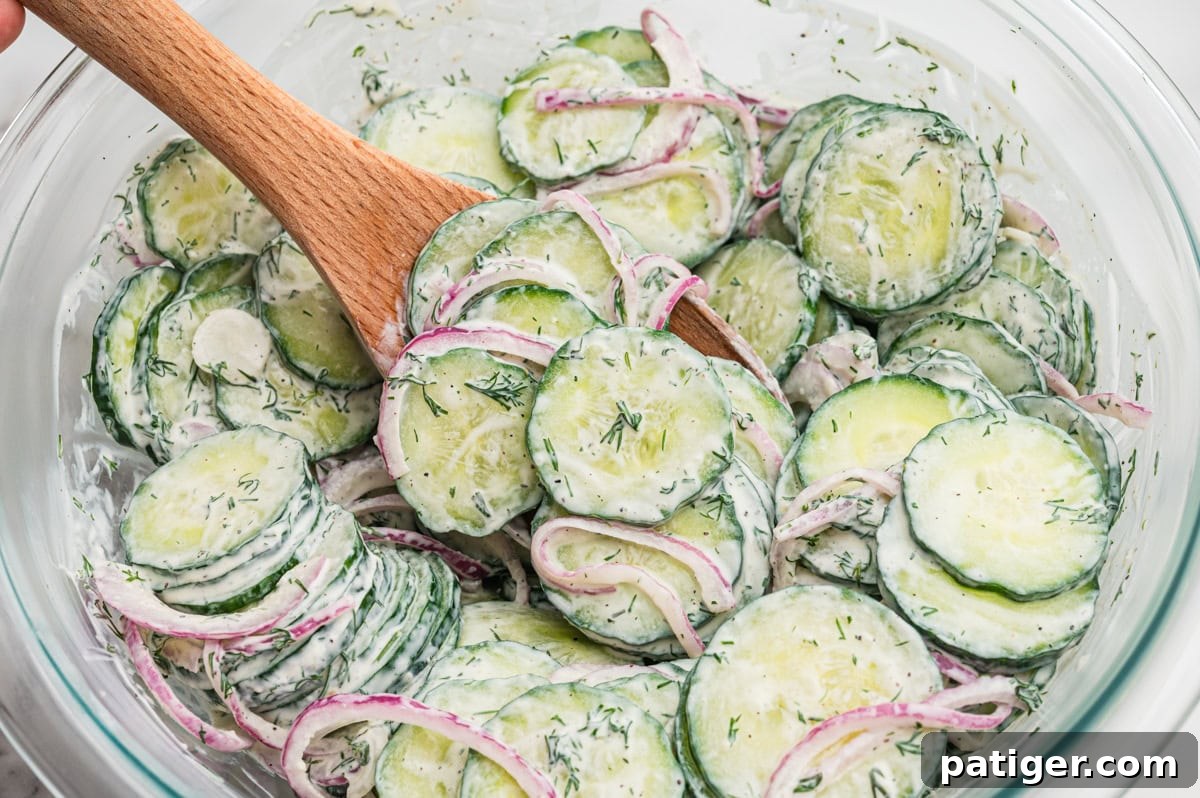 Wooden spoon stirring creamy cucumber salad in a glass bowl, showing cucumber slices, red onion, and creamy dill dressing.