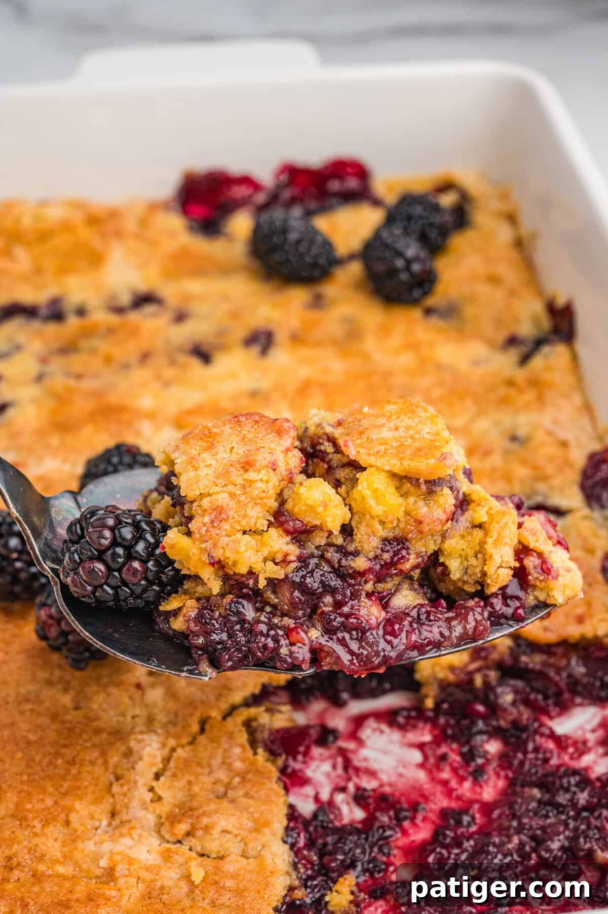 A serving spoon lifting a golden, crumbly scoop of blackberry dump cake, revealing generous amounts of dark berry filling and whole blackberries, over a partially served baking dish.