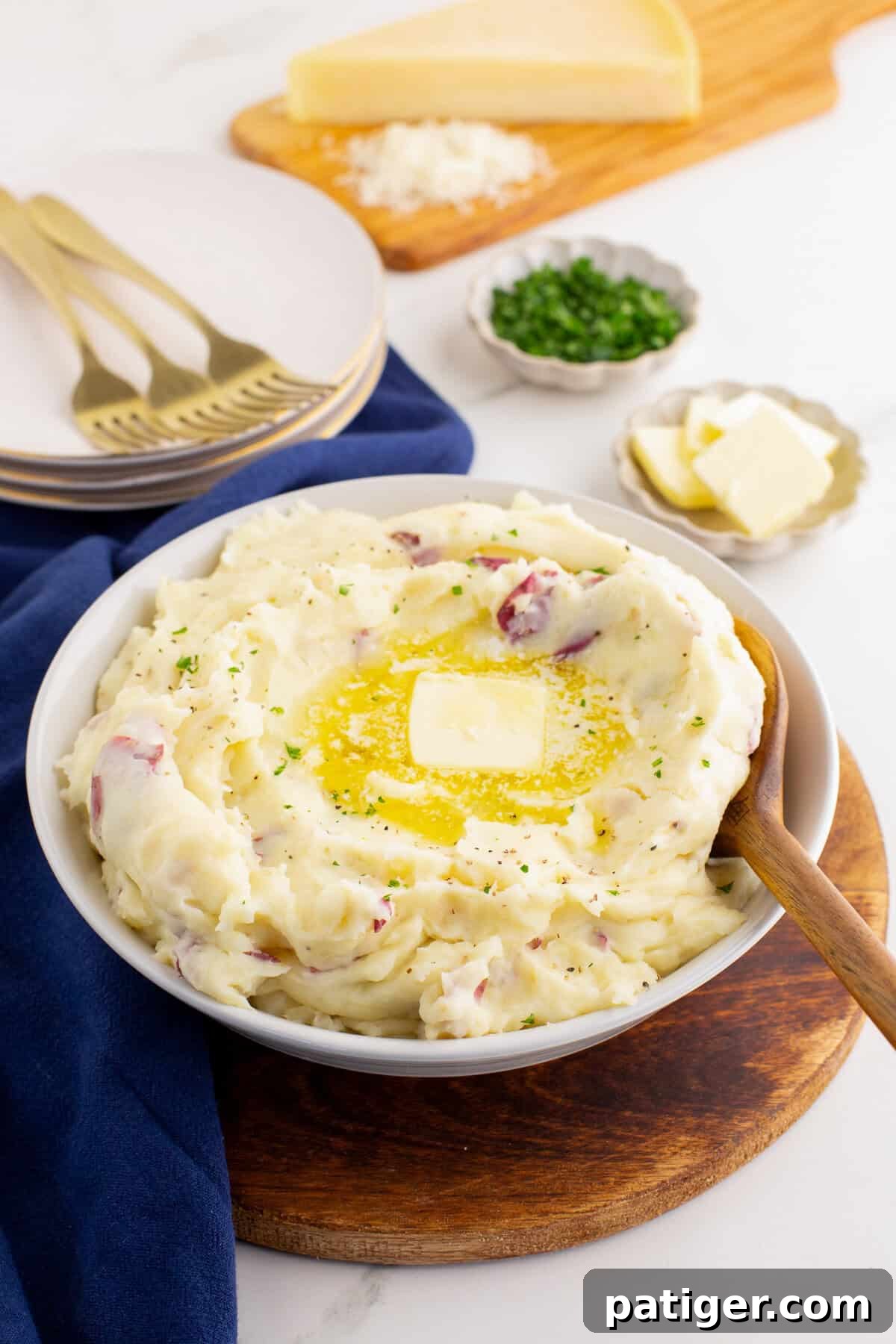 Rustic Red Potato Mash 2 White bowl filled with red skin mashed potatoes, topped with a pat of melting butter and sprinkled with herbs, set on a wooden board with plates, forks, Parmesan cheese wedge, chopped chives, and extra butter in the background.