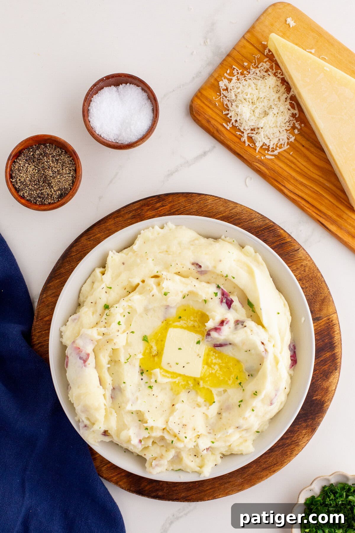 Rustic Red Potato Mash 5 Overhead photo of a white bowl of mashed potatoes with red skin potatoes, garnished with a pat of melting butter and herbs, set on a wooden board with bowls of salt and pepper, a wedge of Parmesan cheese, and shredded cheese on a wooden cutting board.