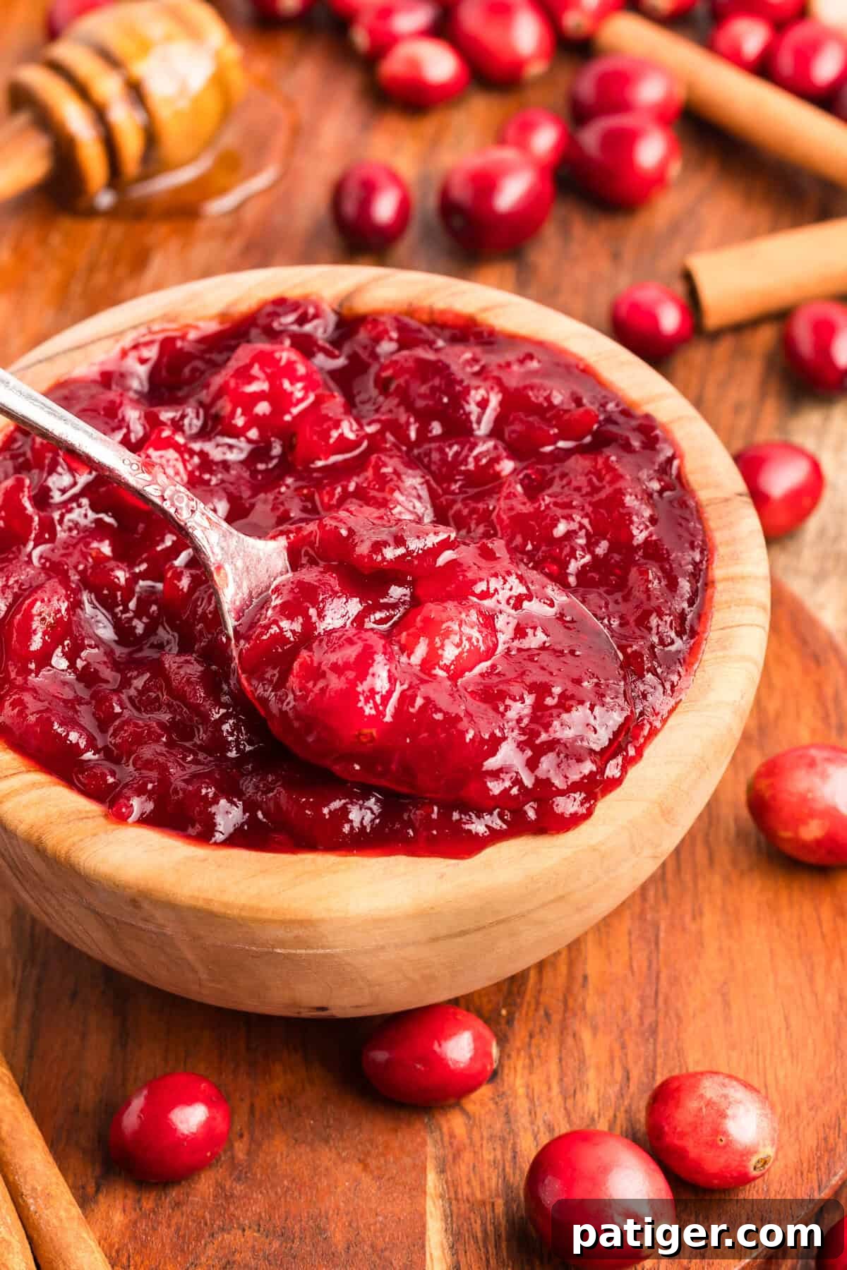 A close-up of a silver spoon lifting a scoop of thick, chunky cranberry sauce from a wooden bowl. The sauce is glossy and vibrant red.