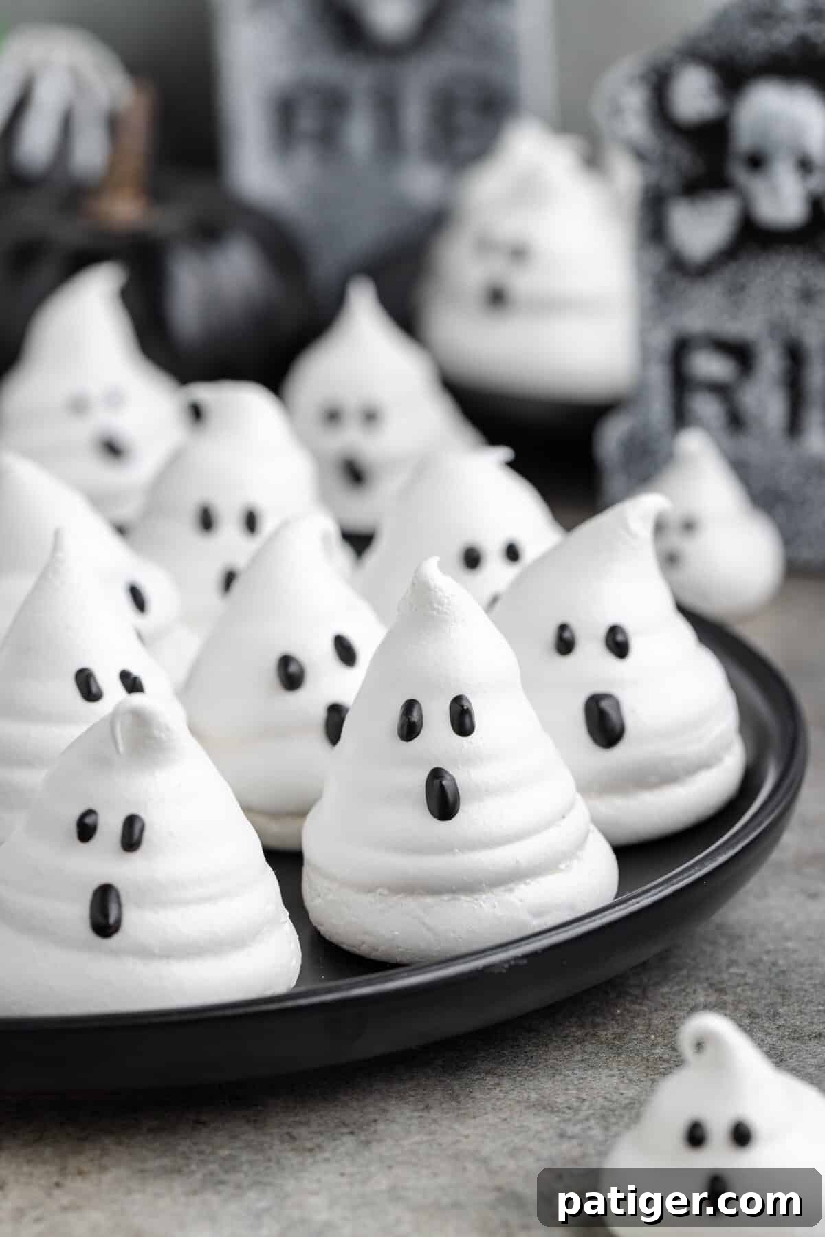 Ghost meringues with black candy eyes and mouths are arranged on a plate. The background includes Halloween-themed tombstone decorations and black pumpkins, creating a festive setting.