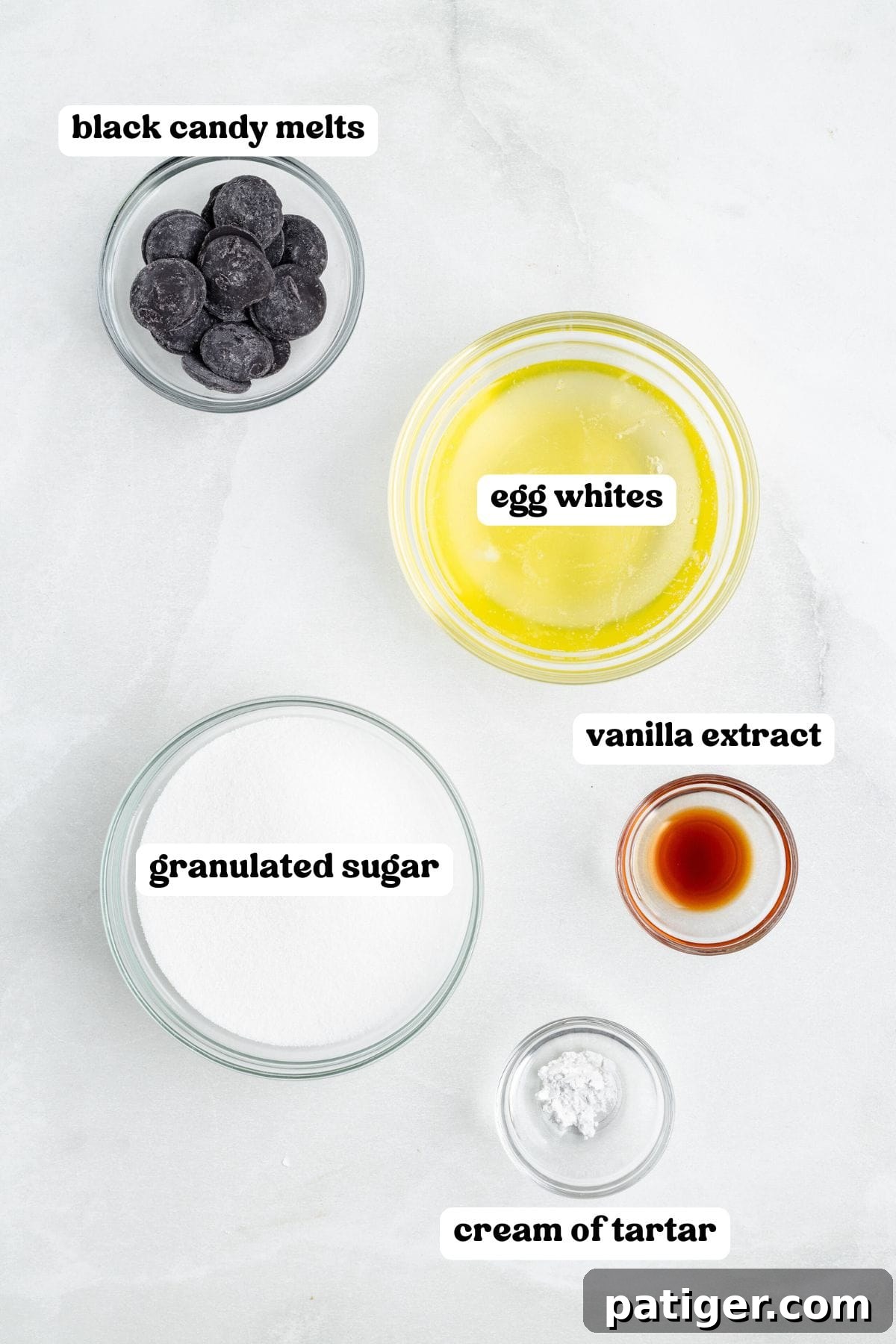 Overhead view of labeled ingredients in clear bowls including: black candy melts, egg whites, granulated sugar, vanilla extract, and cream of tartar.