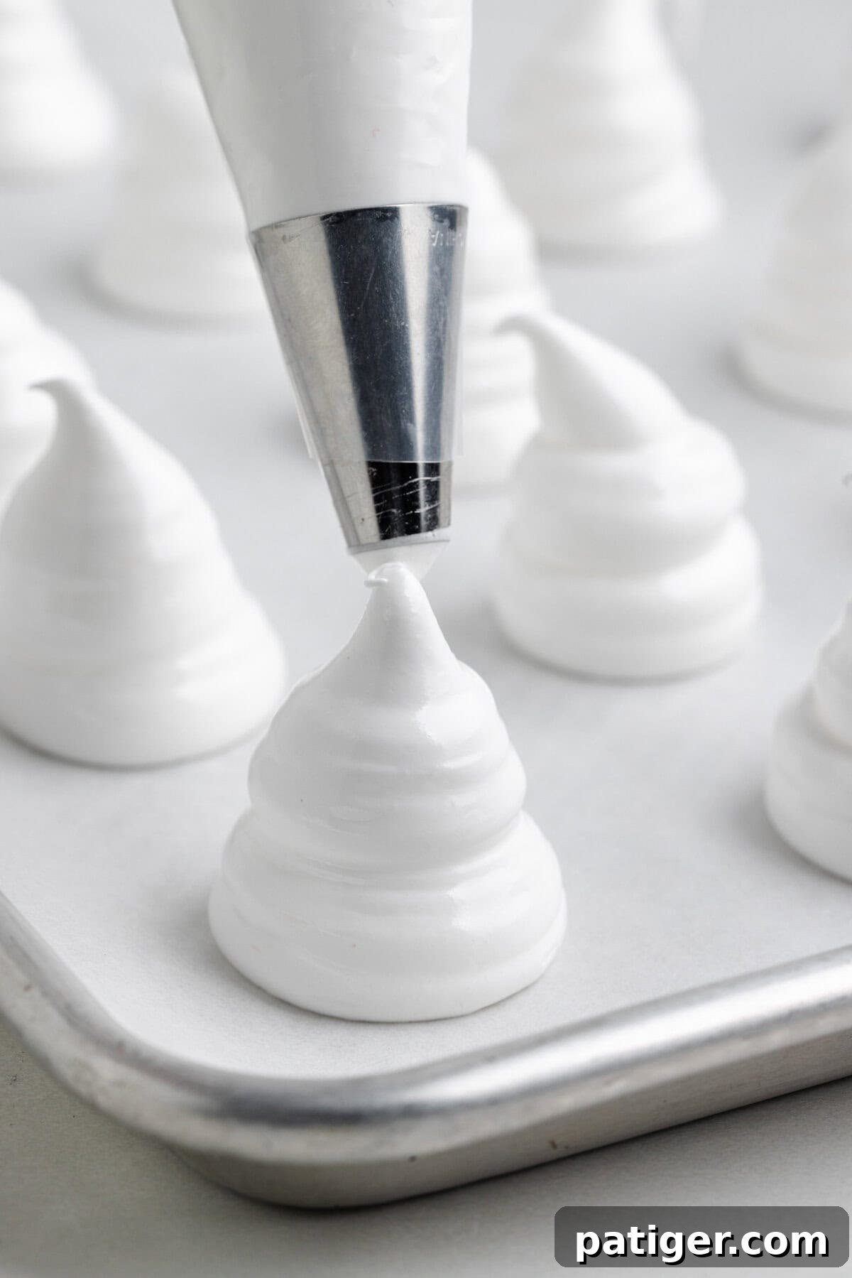 A piping tip forms a peaked swirl of white meringue on a parchment-lined baking tray.