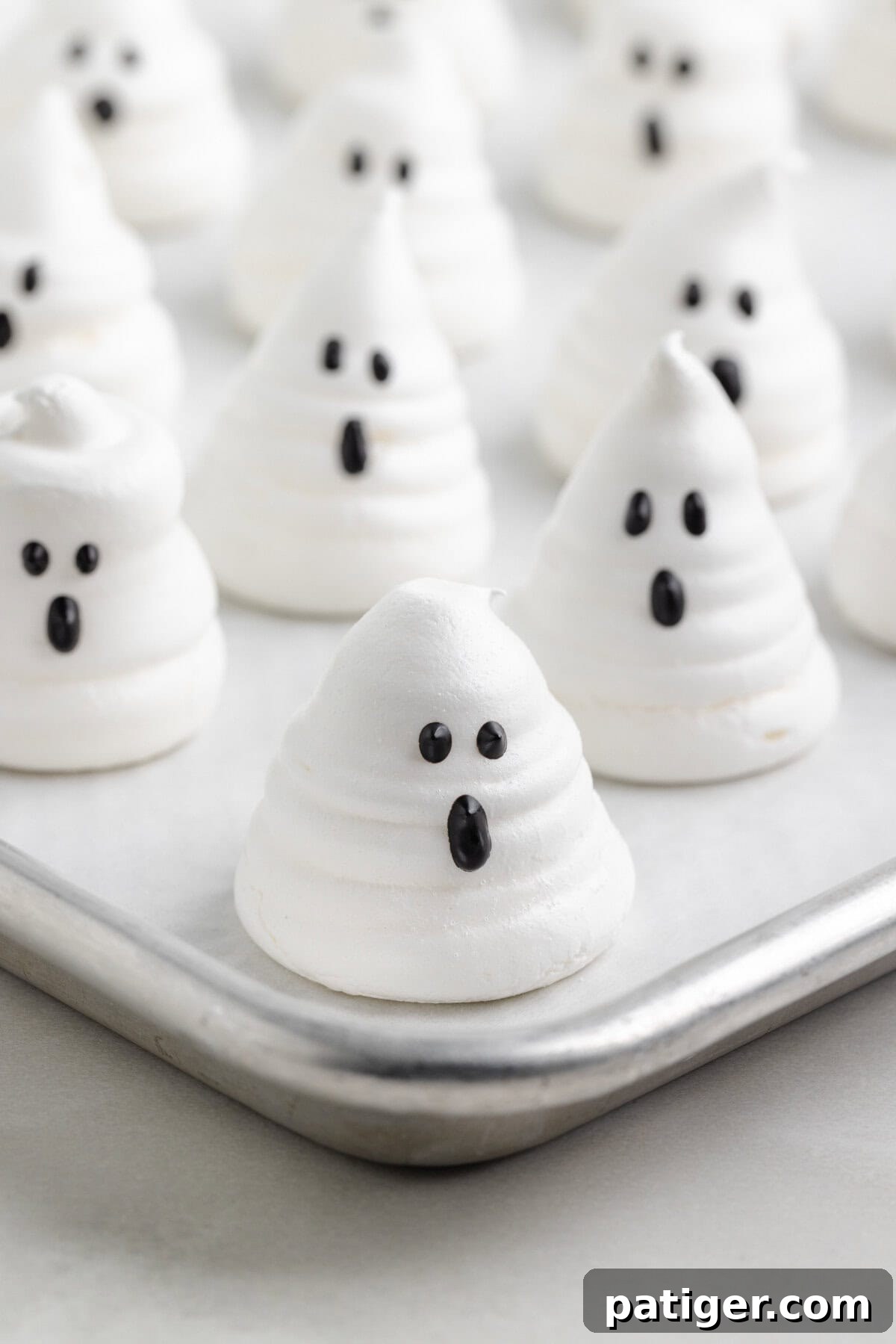 Baked meringue ghosts with black candy melt eyes and oval-shaped mouths arranged on a parchment-lined tray.