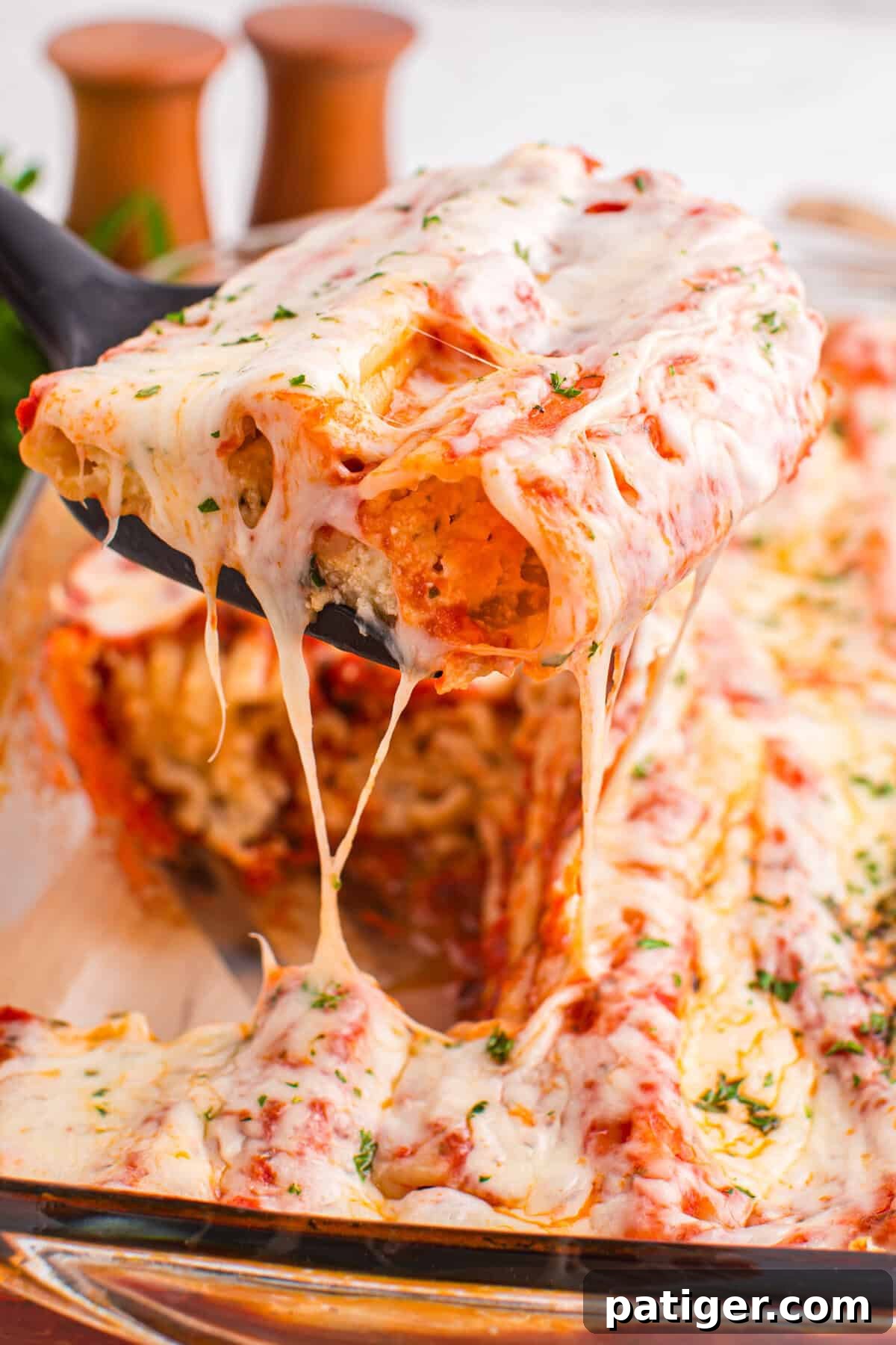 A close-up of a spatula lifting two stuffed manicotti from a baking dish, with stretchy melted cheese and a visible creamy filling inside the pasta. The dish is surrounded by more baked pasta and cheese.