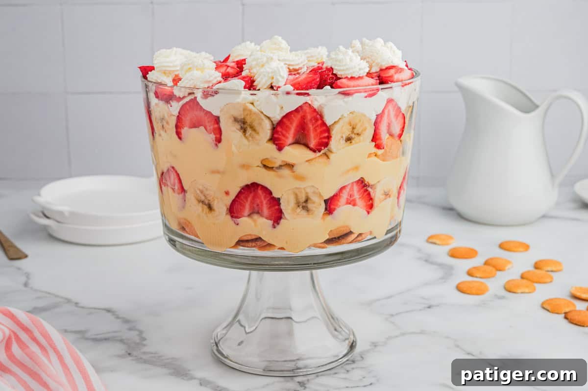  A glass trifle bowl filled with strawberry banana pudding, showing layered rows of sliced strawberries, bananas, vanilla pudding, whipped cream, and Nilla wafers, with whipped cream dollops and strawberries on top. 