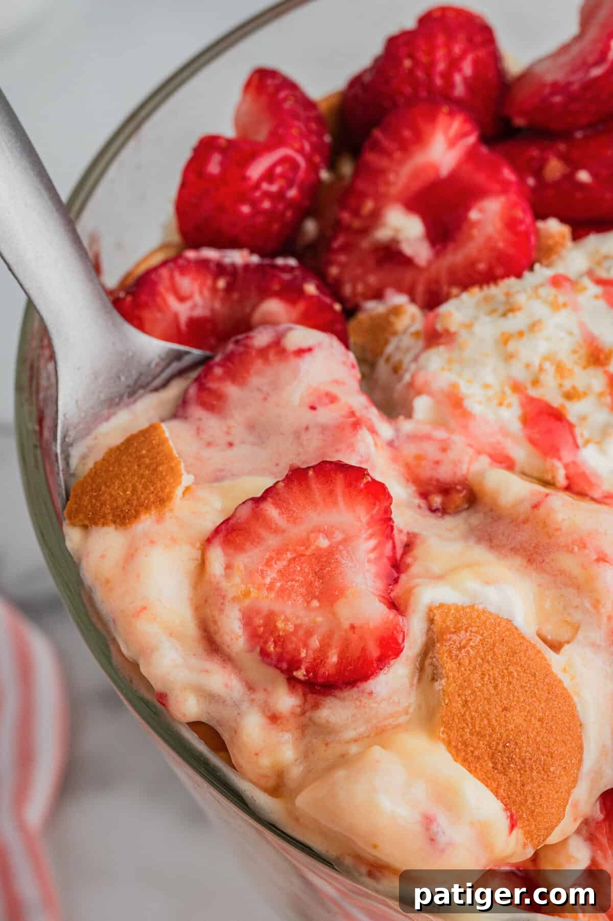 A spoon holding a portion of the strawberry banana pudding dessert with strawberries, whipped cream, pudding, and pieces of Nilla wafers, shown in a close-up view above the trifle bowl.