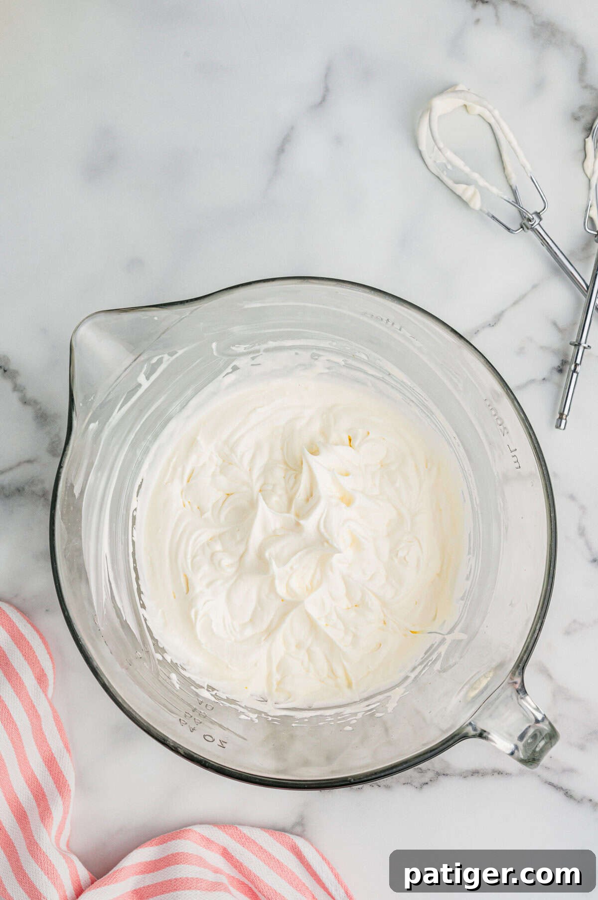 A glass mixing bowl filled with freshly whipped cream on a marble countertop, with electric mixer beaters nearby. 