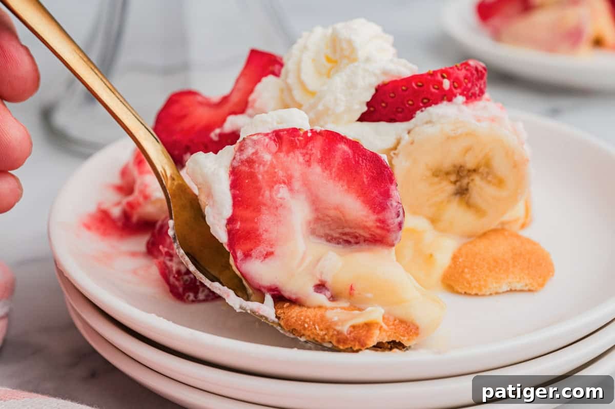 A close-up of a spoon lifting a bite of strawberry banana pudding from a white plate, showing layers of sliced strawberries, banana, vanilla pudding, whipped cream, and a Nilla wafer on the bottom.