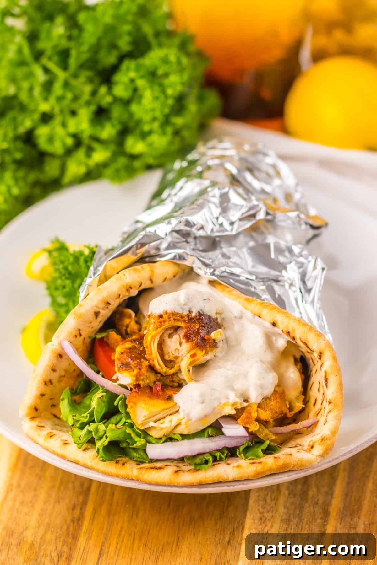 Melt-in-Your-Mouth Slow Cooker Chicken Shawarma 2 A slow cooker chicken shawarma wrap on a white plate, filled with greens, tomato, red onion, shredded chicken, and sauce, wrapped in foil. Garnished with parsley and lemon slices.