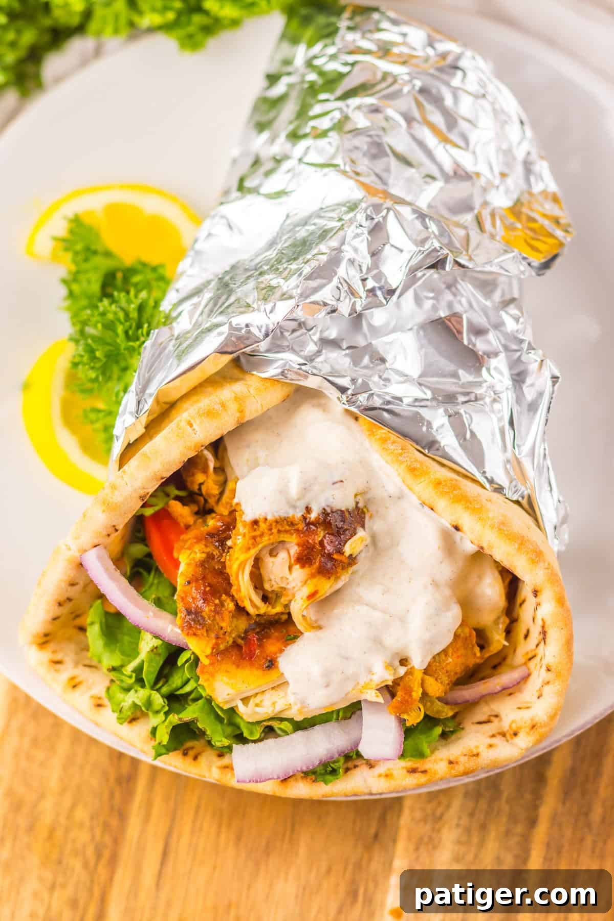Melt-in-Your-Mouth Slow Cooker Chicken Shawarma 9 Slow Cooker chicken shawarma wrap in pita bread, filled with shredded chicken, leafy lettuce, tomato, red onion, and creamy sauce, partially wrapped in foil with lemon slices and parsley on the plate.