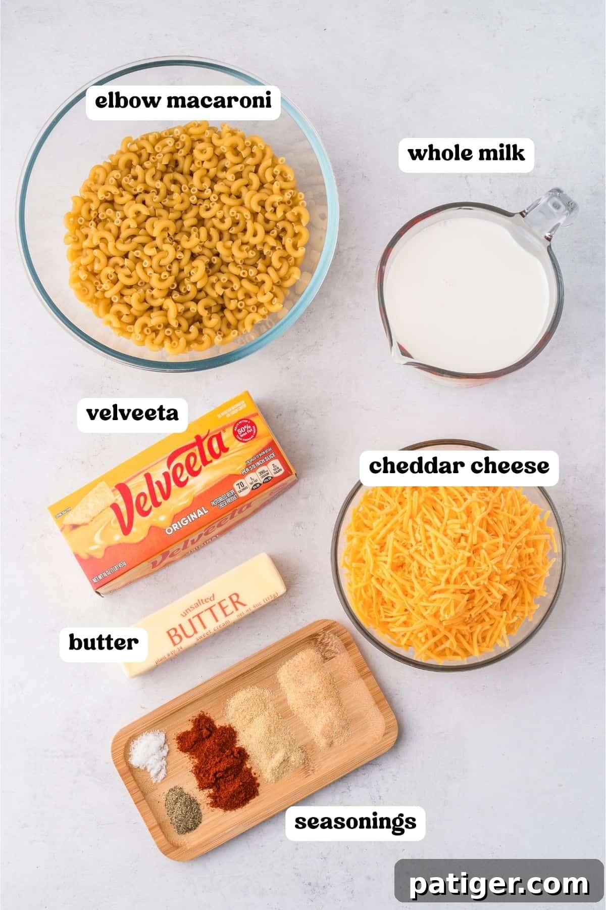 Overhead image displaying clearly labeled ingredients for slow cooker mac and cheese: dry elbow macaroni in a glass bowl, whole milk in a measuring cup, a mound of shredded cheddar cheese, a block of Velveeta cheese, a stick of unsalted butter, and a small wooden tray holding assorted seasonings like garlic powder, onion powder, and paprika.