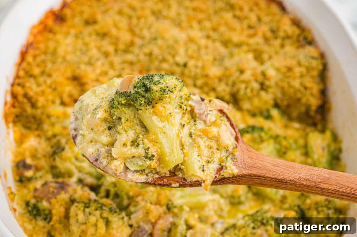 A wooden spoon lifts a creamy portion of broccoli casserole above the baking dish, showing broccoli, mushrooms, and melted cheese and onion beneath a crispy topping.