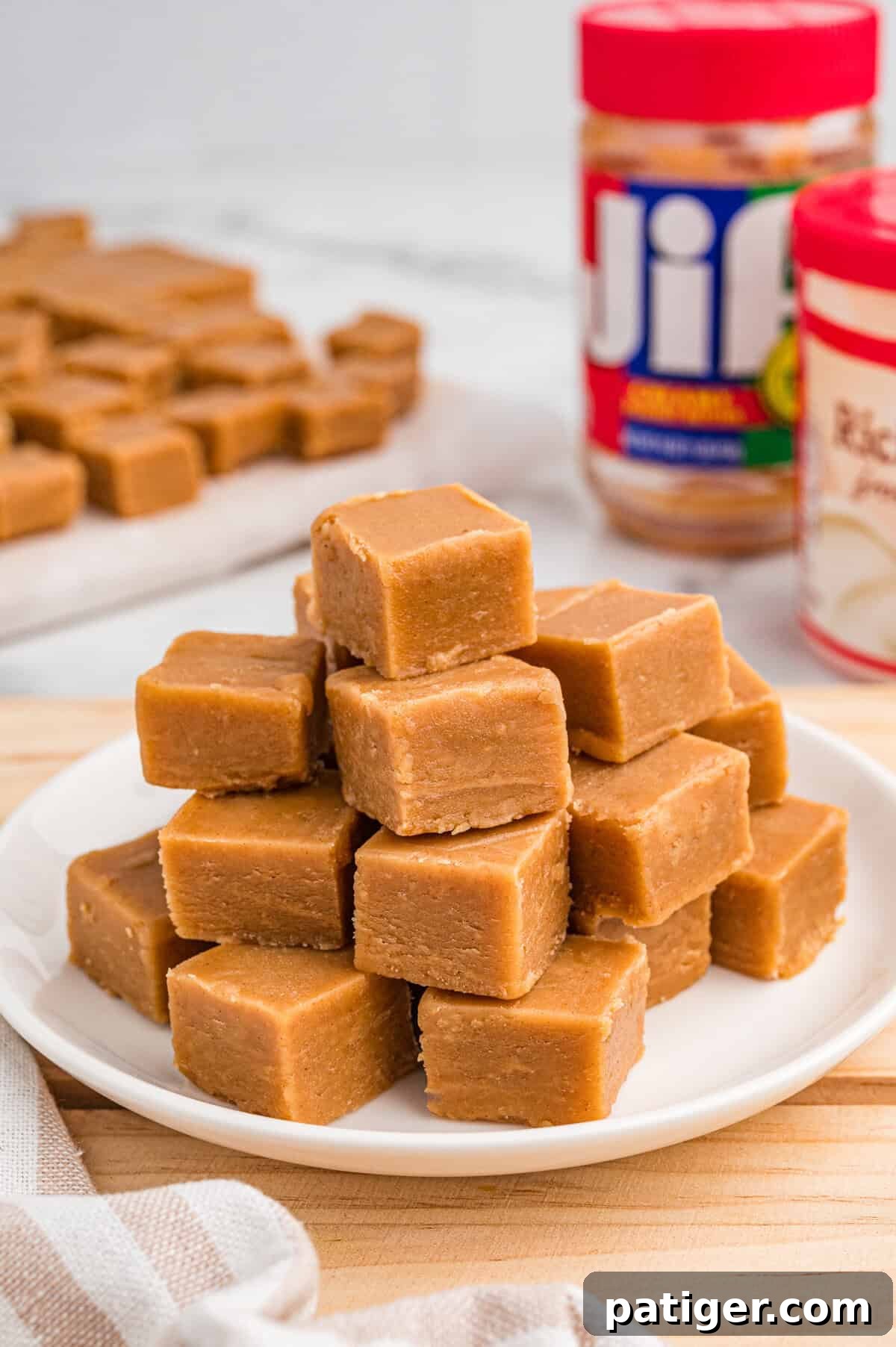 Stack of smooth, light brown peanut butter fudge cubes arranged on a white plate. In the background, jars of Jif peanut butter and Betty Crocker vanilla frosting are slightly blurred, with more fudge pieces visible on parchment paper.