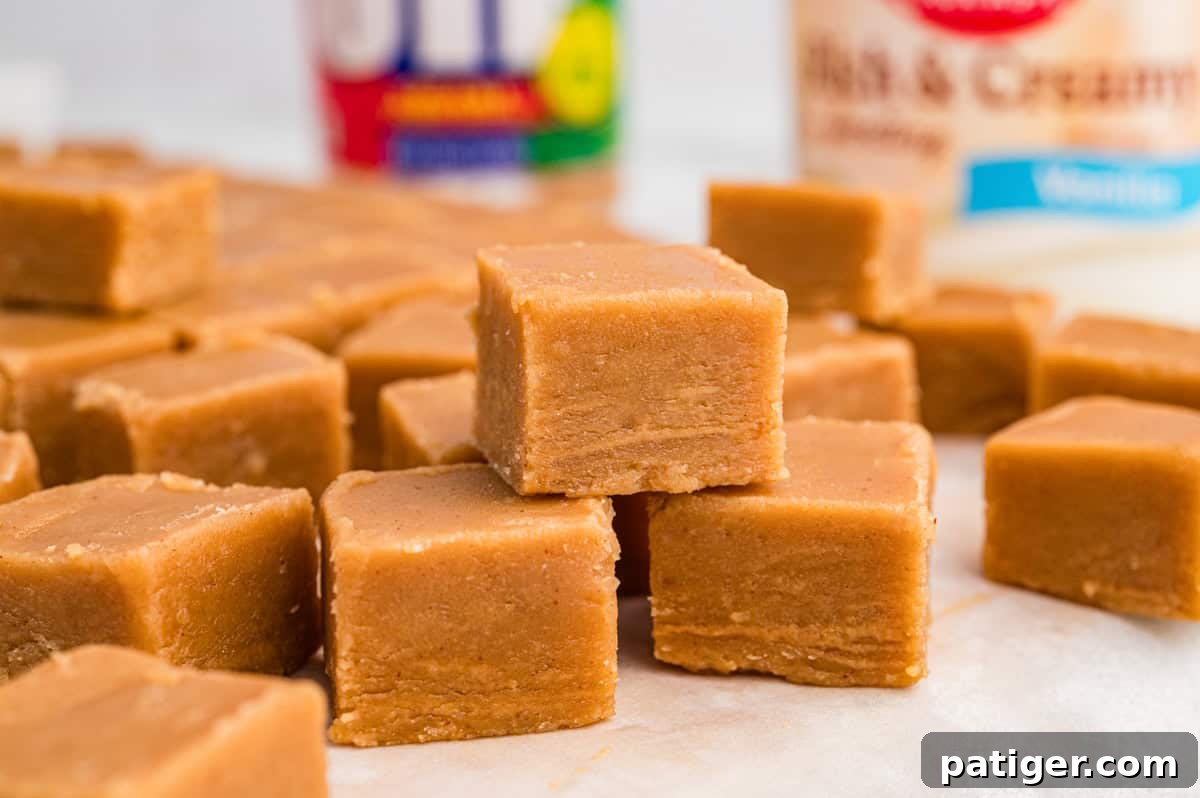 Several cubes of peanut butter fudge resting on parchment paper. A Jif peanut butter jar and a can of frosting are blurred in the background.