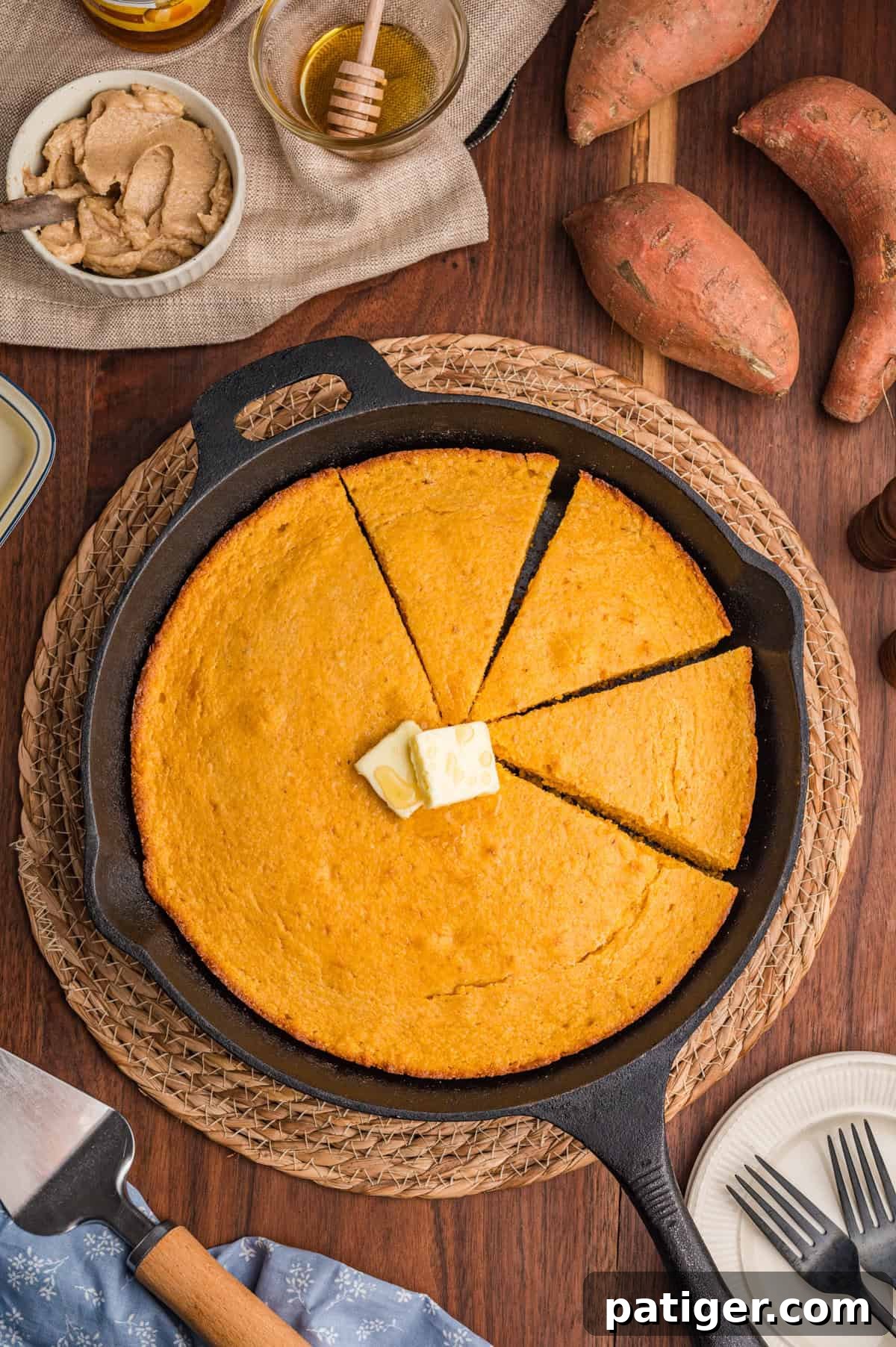 Overhead view of a cast iron skillet filled with sweet potato cornbread, sliced into wedges. A pat of butter is melting in the center, surrounded by sweet potatoes, a honey jar, cinnamon butter, and a serving spatula on a wooden table