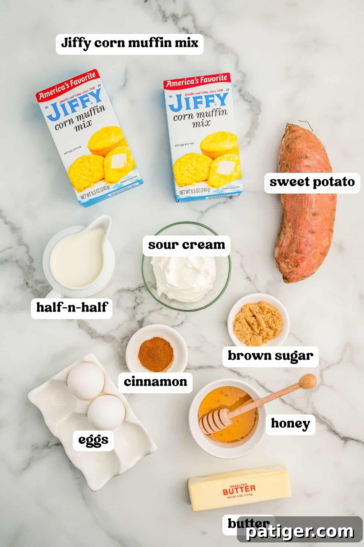 Labeled ingredients for sweet potato cornbread including two boxes of Jiffy corn muffin mix, a sweet potato, sour cream, half-and-half, cinnamon, brown sugar, honey with a dipper, butter, and two eggs.