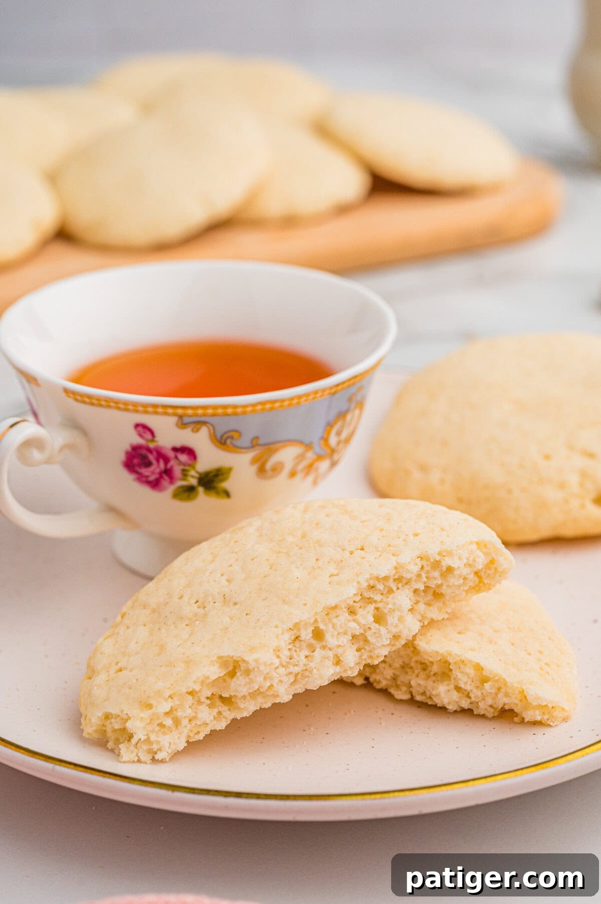 Close-up of a broken old-fashioned tea cake on a ceramic plate, revealing its soft interior. A floral-patterned teacup filled with tea sits behind it, with more cookies in the background.