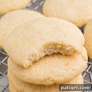 Stacked old fashioned tea cakes with a visible bite out of top cookie revealing a soft, crumbly interior. The cookie sits on a cooling rack with more cookies in the background.