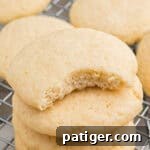 Stacked old fashioned tea cakes with a visible bite out of top cookie revealing a soft, crumbly interior. The cookie sits on a cooling rack with more cookies in the background.