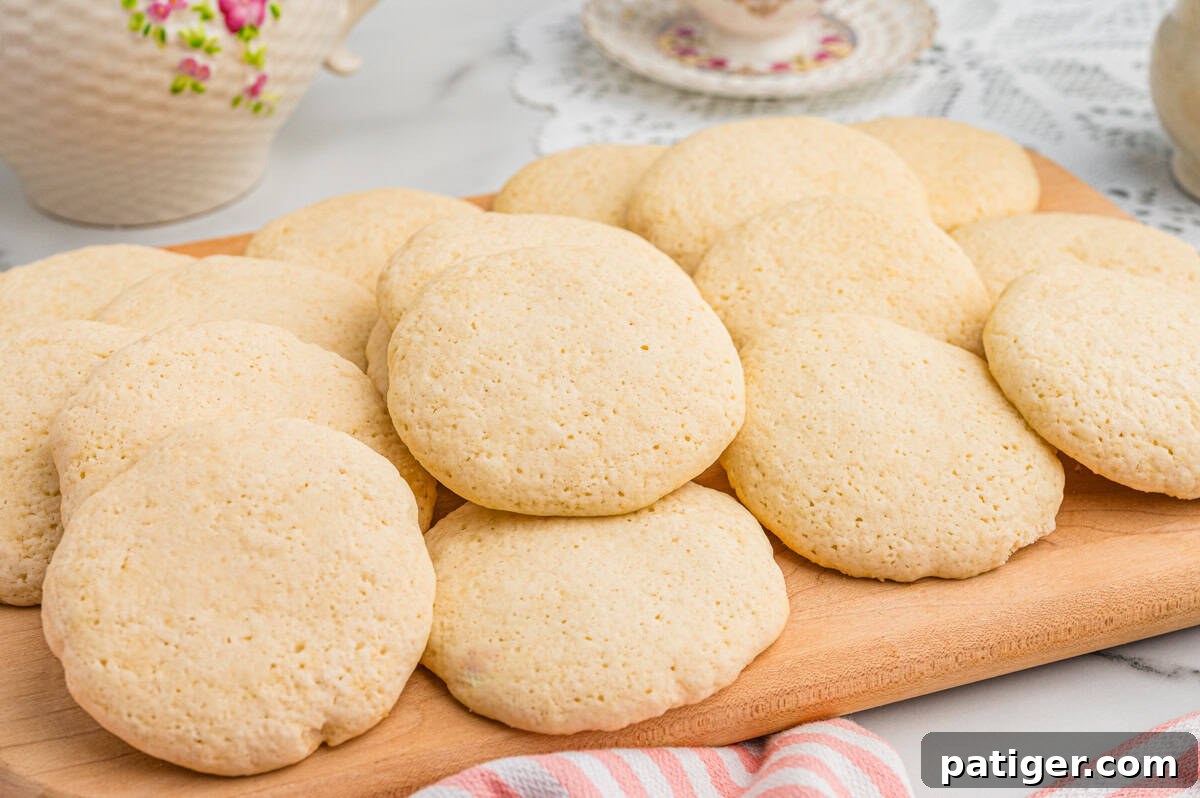 Angled view of old-fashioned tea cakes stacked and layered on a wooden board. A floral teapot and teacup sit in the background on a lace doily, suggesting a tea-time setting.