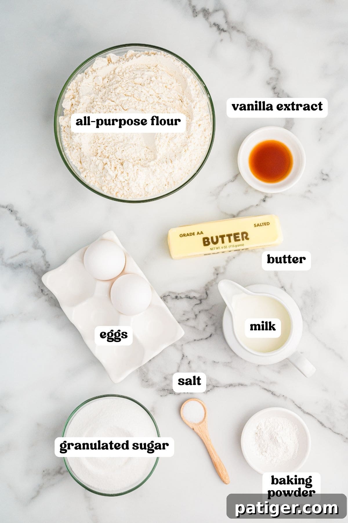Labeled ingredient for making old-fashioned tea cakes. Items include a bowl of all-purpose flour, two eggs in a ceramic holder, granulated sugar, a stick of salted butter, a small bowl of vanilla extract, milk in a white pitcher, salt in a wooden spoon, and baking powder in a small bowl.
