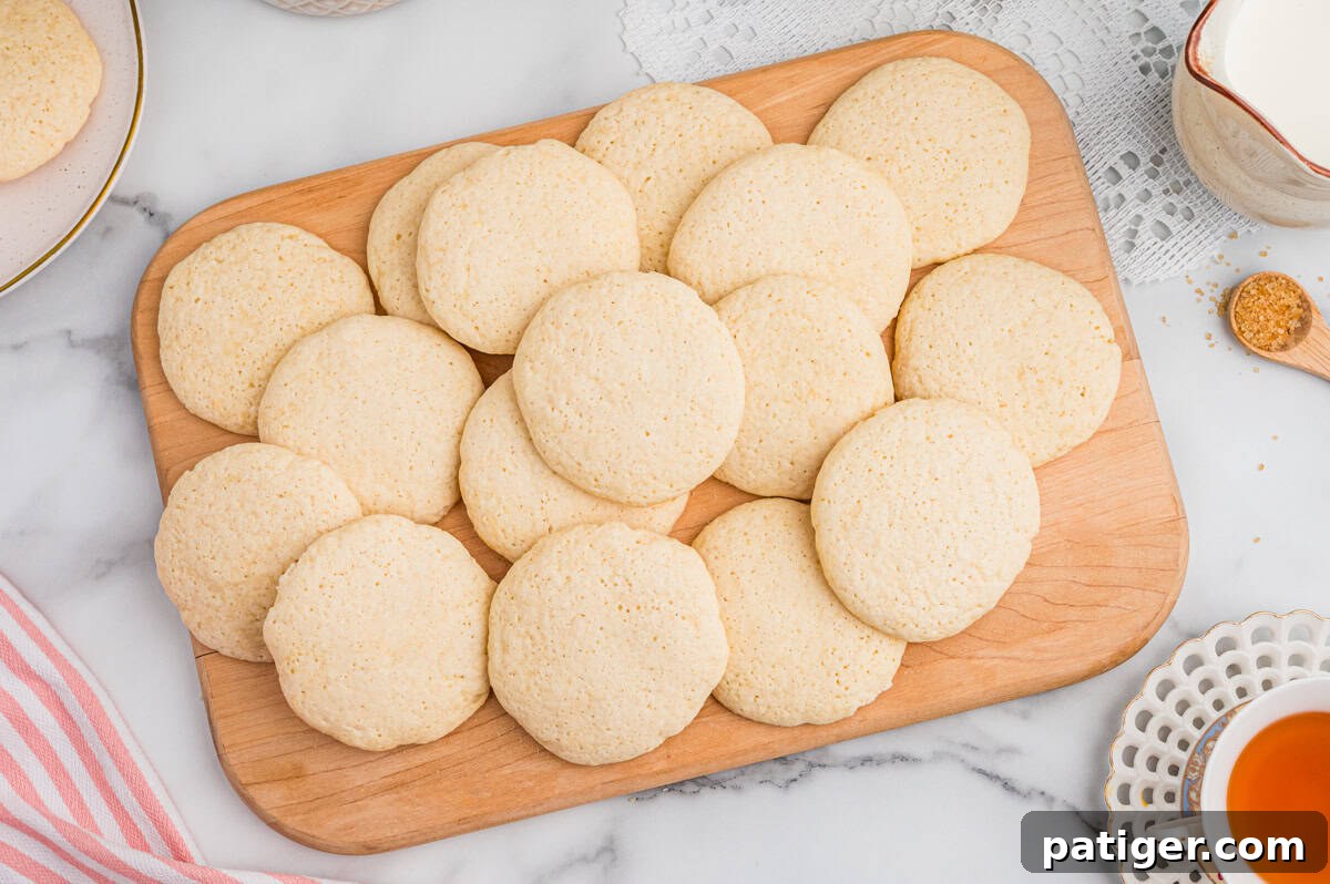 Overhead view of a batch of old-fashioned tea cakes arranged on a rectangular wooden board. The cookies are pale golden and round, placed on a marble surface with lace and ceramic dishes nearby.