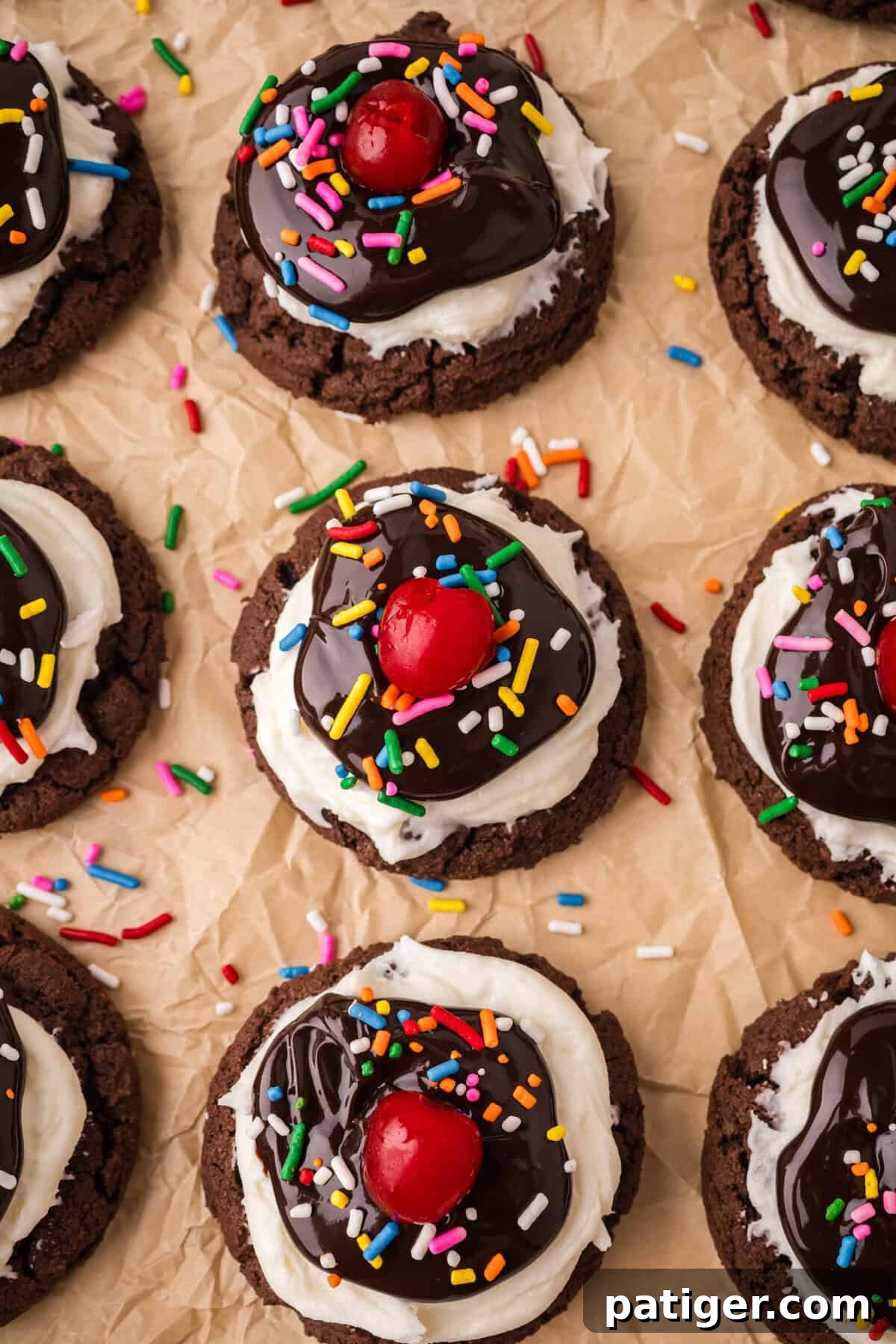 A top-down arrangement of freshly decorated Brownie Sundae Cookies on parchment paper, each showcasing a generous dollop of white frosting, a swirl of chocolate sauce, a scattering of colorful rainbow sprinkles, and a bright red maraschino cherry on top, with extra sprinkles loosely scattered.