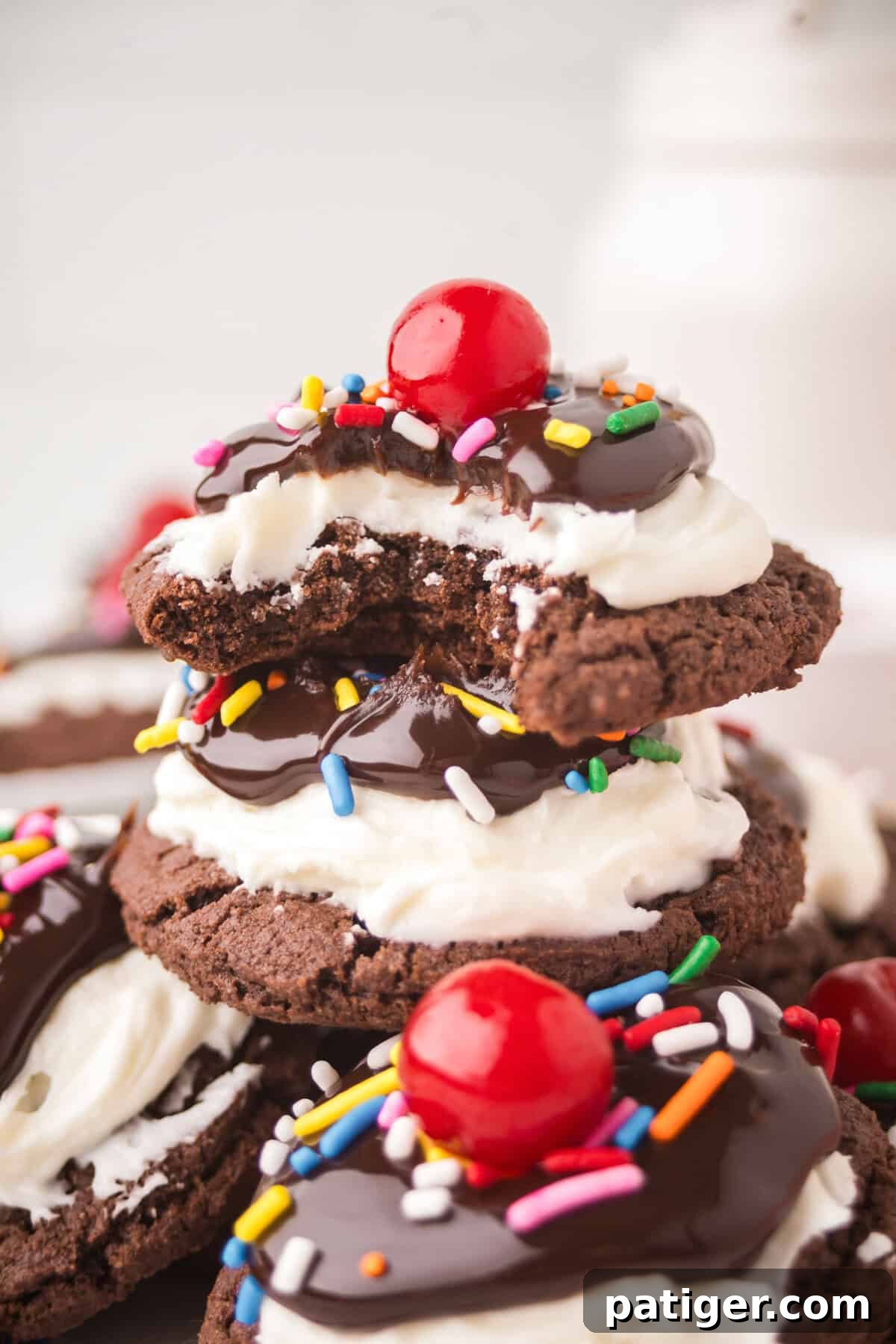 Stacked Ice Cream Sundae Brownie Cookies, richly layered with creamy white frosting, glossy chocolate fudge, colorful sprinkles, and topped with bright red cherries. The top cookie has a delicious bite taken out.