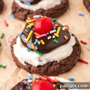 Hot fudge sundae cookie with a chocolate cookie base topped with white frosting, hot fudge sauce, rainbow sprinkles, and a maraschino cherry.