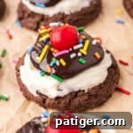 Hot fudge sundae cookie with a chocolate cookie base topped with white frosting, hot fudge sauce, rainbow sprinkles, and a maraschino cherry.