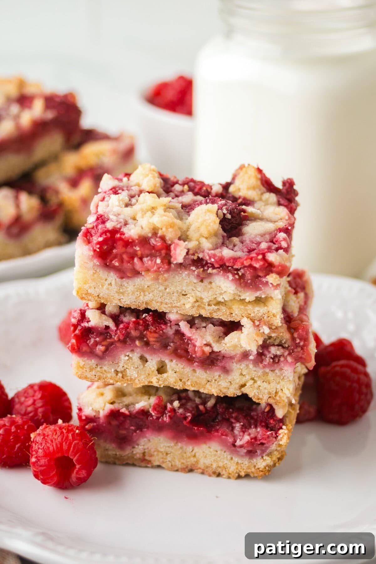 Berry Burst Crumb Bars 2 stack of three raspberry crumble bars on a white plate, next to a glass jar of milk. The bars have golden crusts, thick raspberry layers, and crumbly toppings. Fresh raspberries are scattered around the plate.