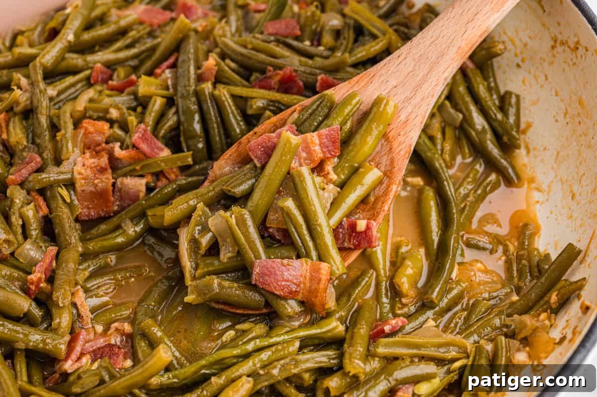 Close-up of green beans and bacon in a shallow pan with broth, being stirred by a wooden spoon.