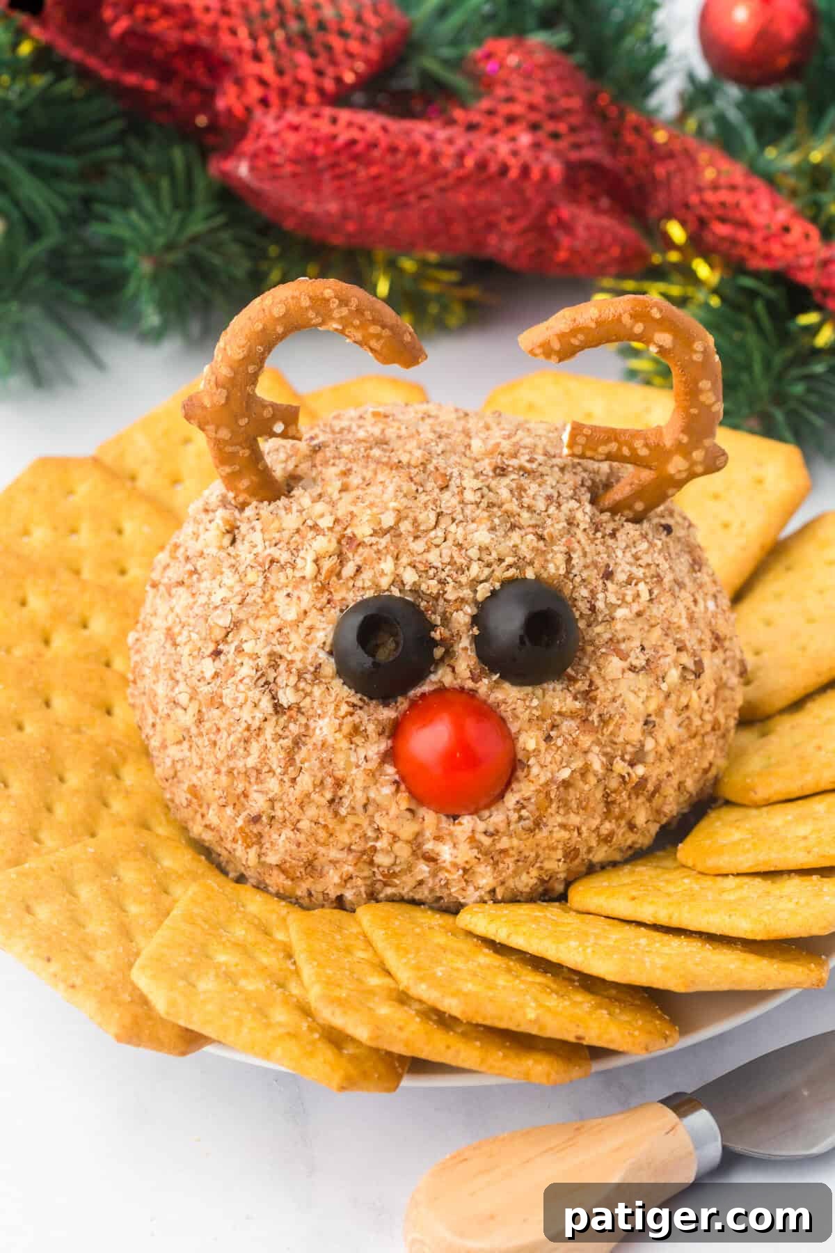 A festive reindeer cheese ball decorated with pretzel antlers, black olive eyes, and a red cherry tomato nose. The cheese ball is coated in chopped nuts and surrounded by golden crackers on a white plate with holiday decorations in the background.