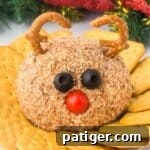 A reindeer cheese ball coated in chopped nuts, with black olives for eyes, a cherry tomato for a nose, and pretzel twists shaped into antlers. The cheese ball is on a plate surrounded by crackers, with holiday decorations in the background.