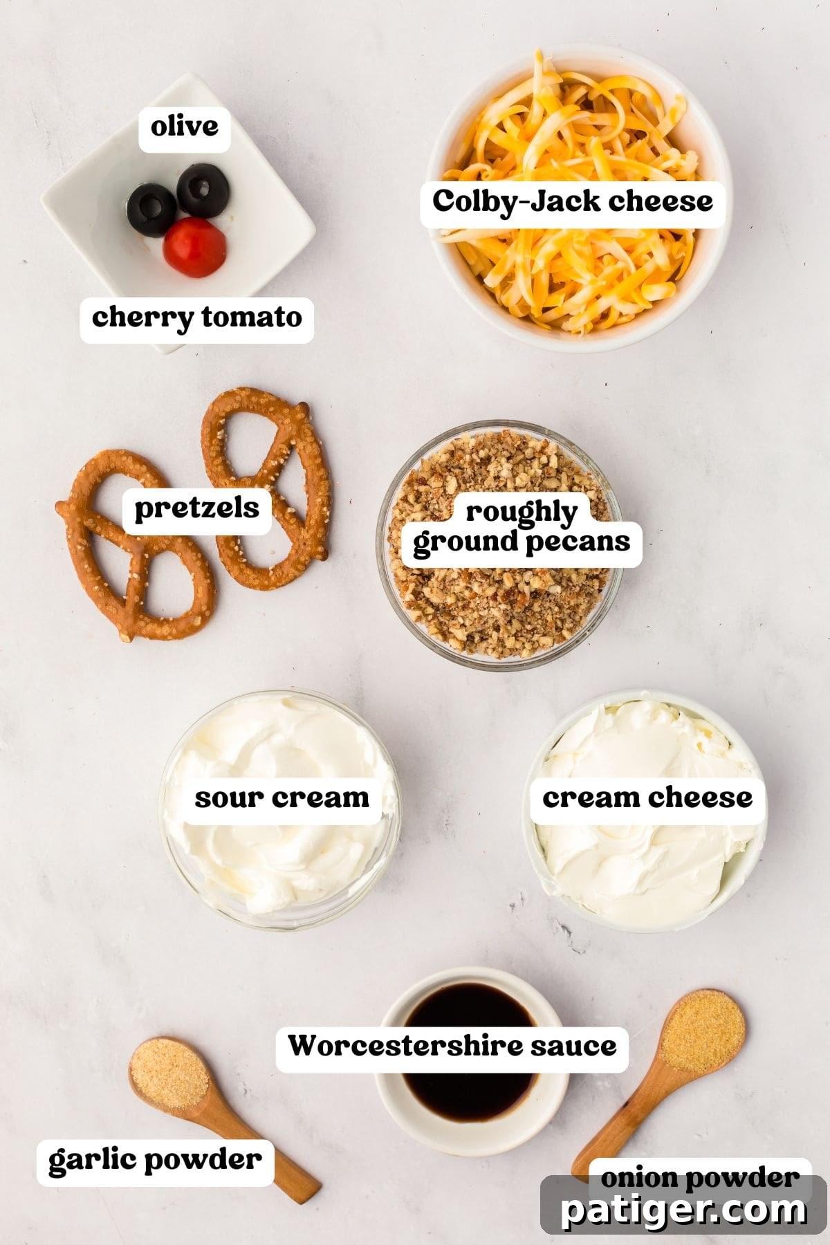 Overhead view of labeled ingredients for a reindeer cheese ball including black olives, a cherry tomato, shredded Colby-Jack cheese, two pretzels, roughly ground pecans, sour cream, cream cheese, Worcestershire sauce, garlic powder, and onion powder.