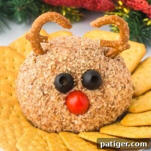 A reindeer cheese ball coated in chopped nuts, with black olives for eyes, a cherry tomato for a nose, and pretzel twists shaped into antlers. The cheese ball is on a plate surrounded by crackers, with holiday decorations in the background.