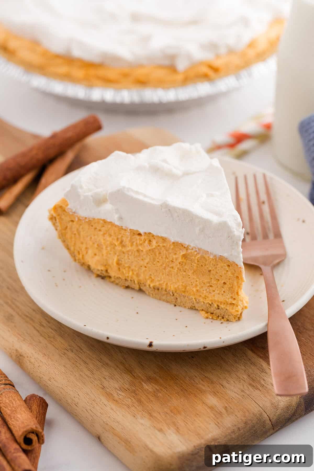 Velvety No-Bake Pumpkin Pie Bliss 2 A perfectly sliced no-bake pumpkin pie, showcasing its smooth, creamy filling and golden graham cracker crust, topped with a generous swirl of cinnamon whipped cream, resting on a white plate with a bronze fork.