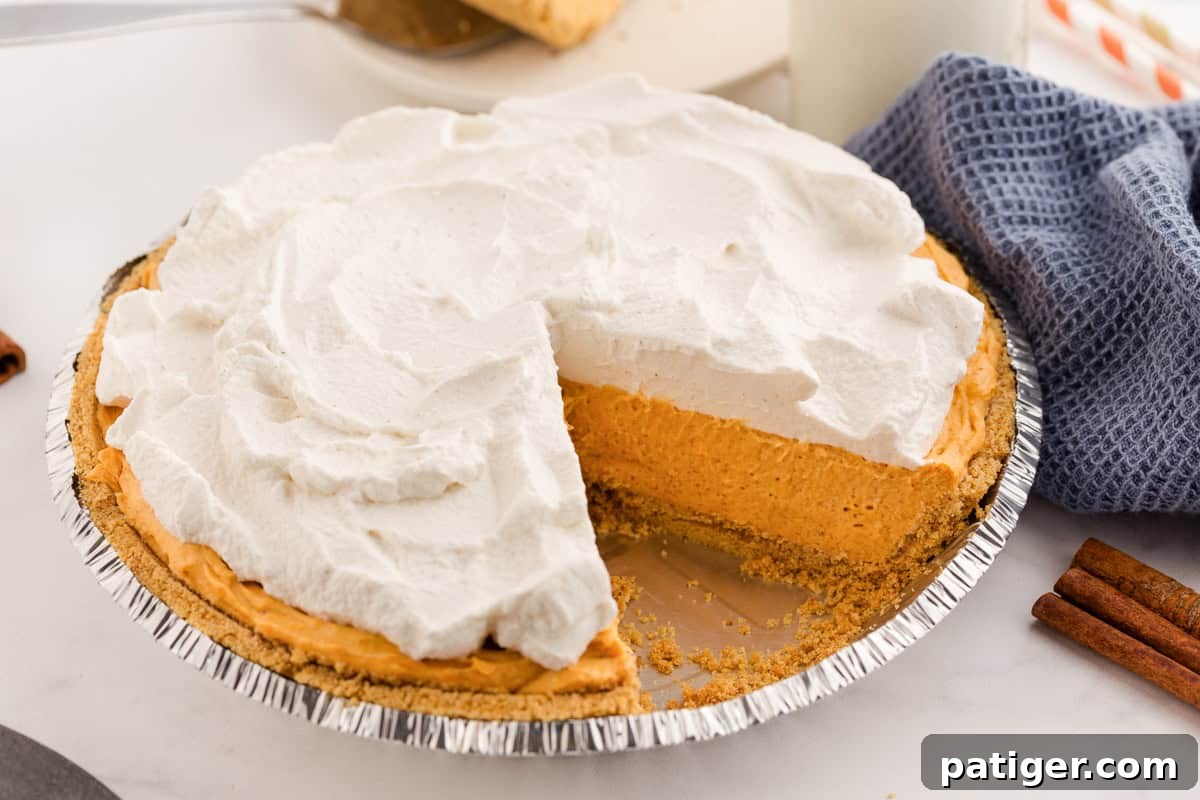 Velvety No-Bake Pumpkin Pie Bliss 3 A full no-bake pumpkin pie in a foil pan, featuring a crisp graham cracker crust and a generous layer of fluffy whipped topping. A single slice has been removed, revealing the inviting, creamy pumpkin filling beneath.