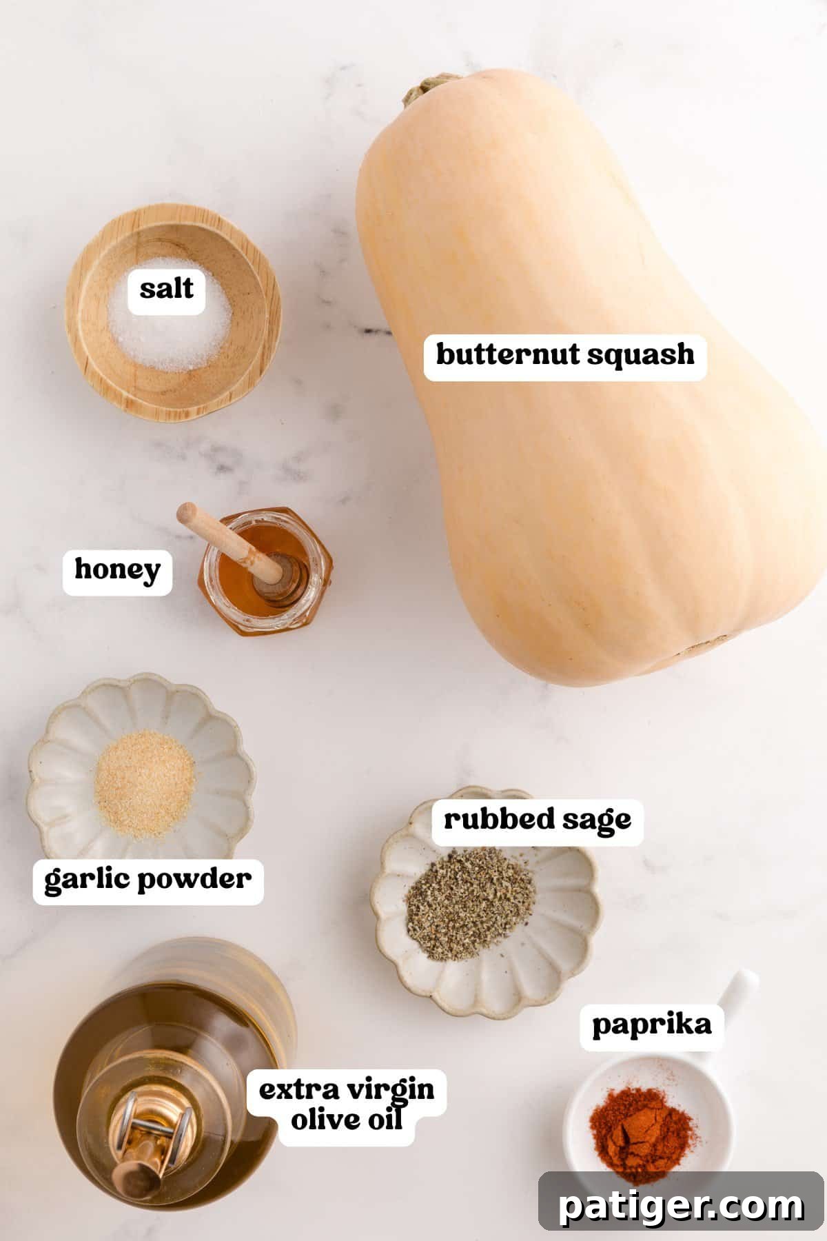 Labeled ingredients for roasted butternut squash, including a whole butternut squash, salt, honey, garlic powder, rubbed sage, paprika, and a bottle of extra virgin olive oil, laid out on a white surface.
