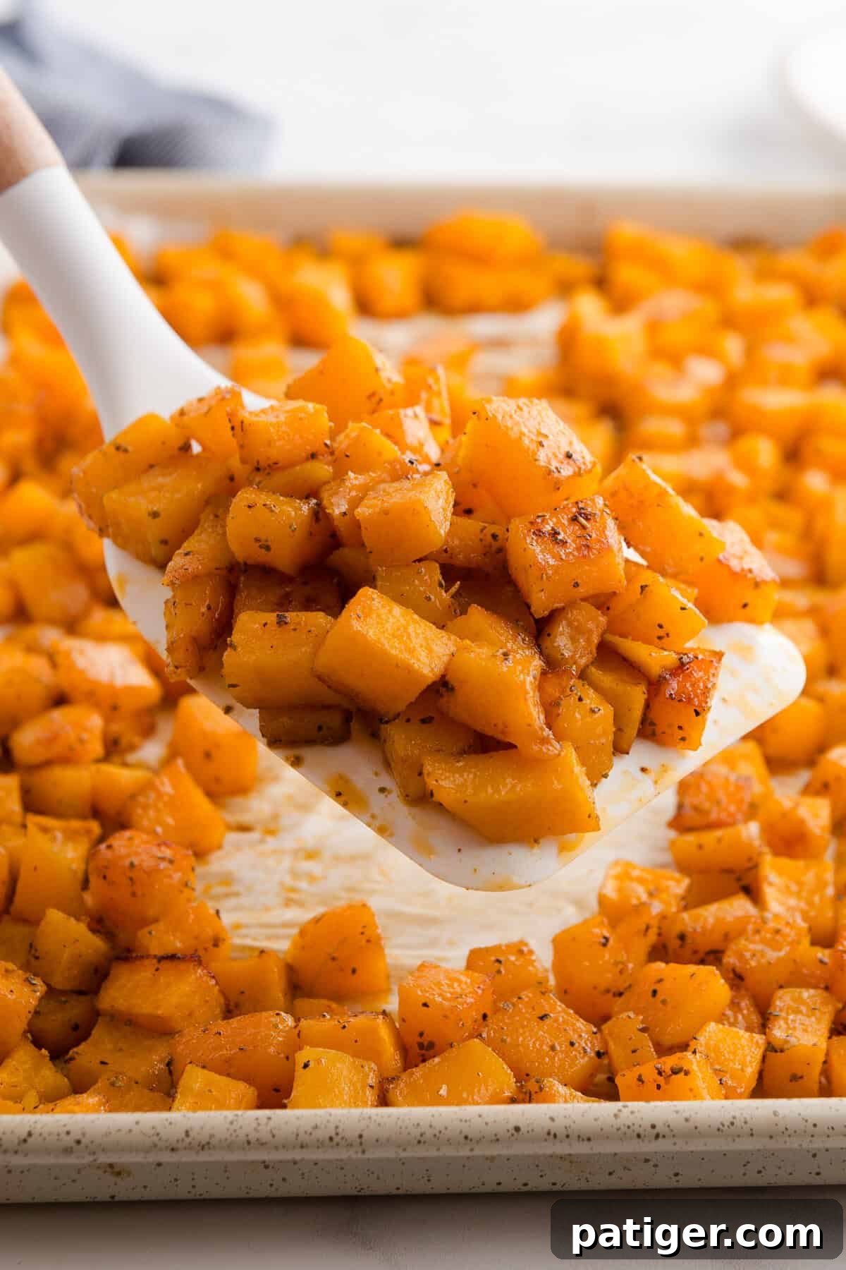 A spatula lifting a portion of roasted butternut squash cubes from a parchment-lined baking sheet. The squash is golden and seasoned, with the rest of the tray visible in the background, highlighting its perfect roast.