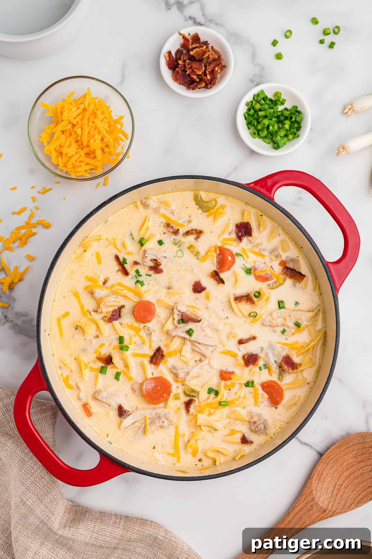 Overhead image of crack chicken noodle soup in a Dutch oven. Surrounding the pot are bowls of shredded cheddar, crumbled bacon, and chopped green onions, and whole green onions.