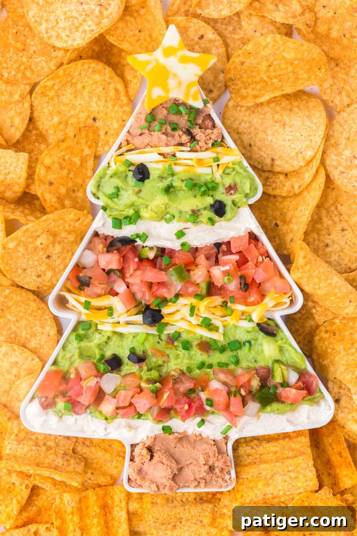 Overhead view of a full Christmas tree-shaped serving dish containing layered taco dip with refried beans, guacamole, sour cream, shredded cheese, diced tomatoes, olives, and green onions. A cheese star decorates the top, with tortilla chips surrounding the dish.