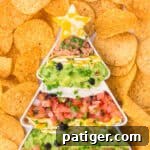 A close-up top view of a Christmas tree-shaped dish filled with layered taco dip, including refried beans, guacamole, sour cream, shredded cheese, diced tomatoes, olives, and green onions. A marbled cheese star tops the tree, and the dish is surrounded by round tortilla chips.