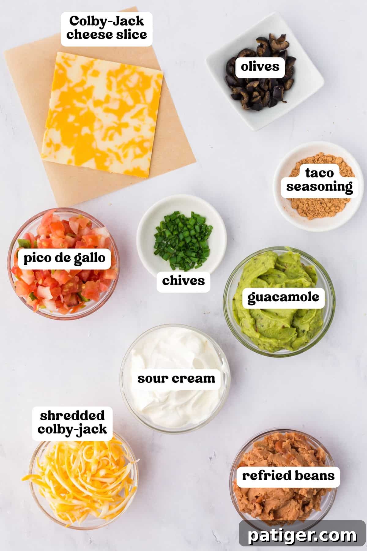 Overhead image of taco dip ingredients in small bowls, each labeled with text. Items include Colby-Jack cheese slice, olives, taco seasoning, chives, guacamole, sour cream, shredded Colby-Jack, pico de gallo, and refried beans.